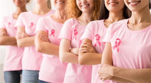 Six women with pink shirts and breast cancer awareness ribbons stand with their arms folded, smiling at the camera. 