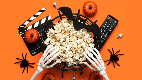 Plastic skeleton hands reaching into a bowl of popcorn with a remote, spiders, pumpkins and a movie clap board surrounding it.