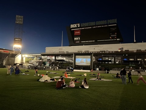 People sitting on the pitch at Dick's Sporting Goods Park watching a movie on a screen. 
